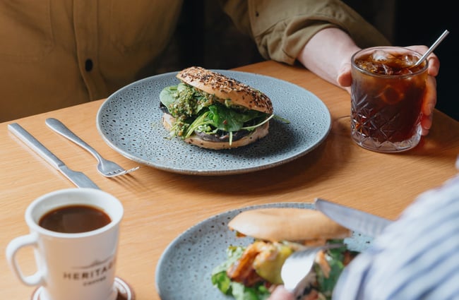 A bagel filled with mushrooms and greens, and an iced coffee on a table.