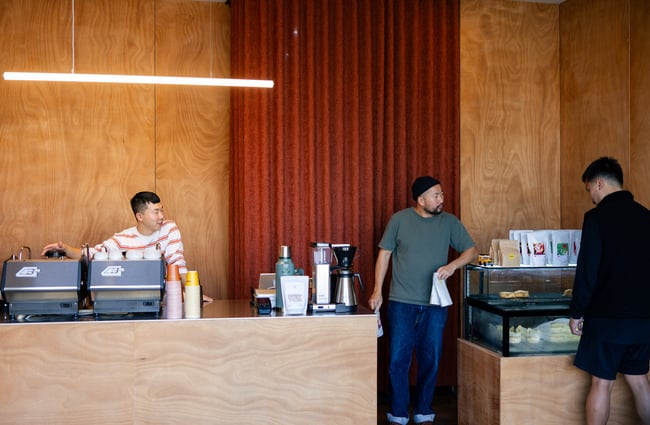 Overall view of Holiday cafe with one barista standing behind the coffee machine and another serving a customer by the food cabinet