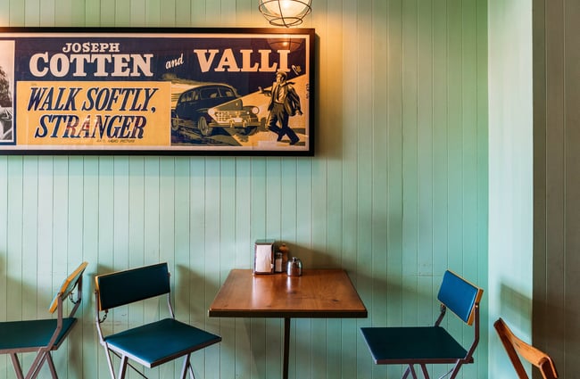 Table and two chairs against a mint-green wall inside a cafe with a vintage poster on the wall