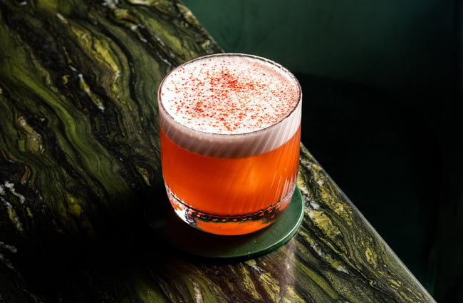 Close up of a pink-hued, foamy-topped cocktail in a lowball glass sitting on a green marble counter.