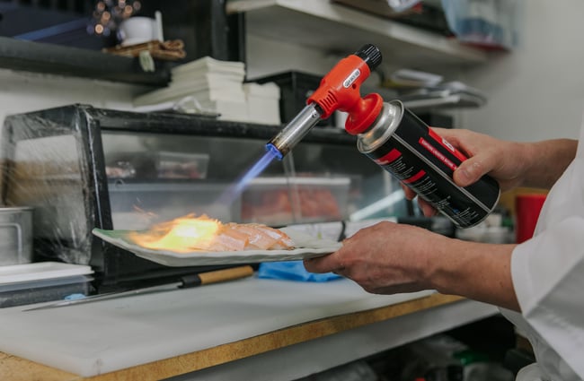 A Kinji Chef using a blow torch to add the final touches to a plate of sushi.