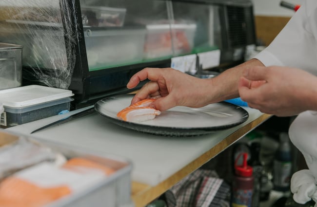 A Chef using his hands to place a piece of salmon sushi on a plate.