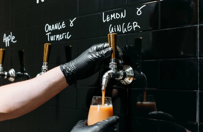 A worker in black gloves pouring lemon and ginger kombucha from the tap.