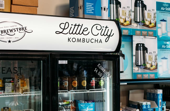 A drinks fridge of with a black and white Little City Kombucha label.