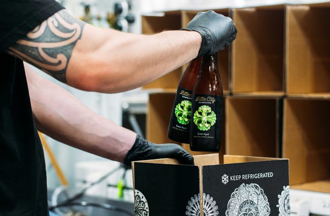 A worker wearing black gloves putting bottles of kombucha into boxes.