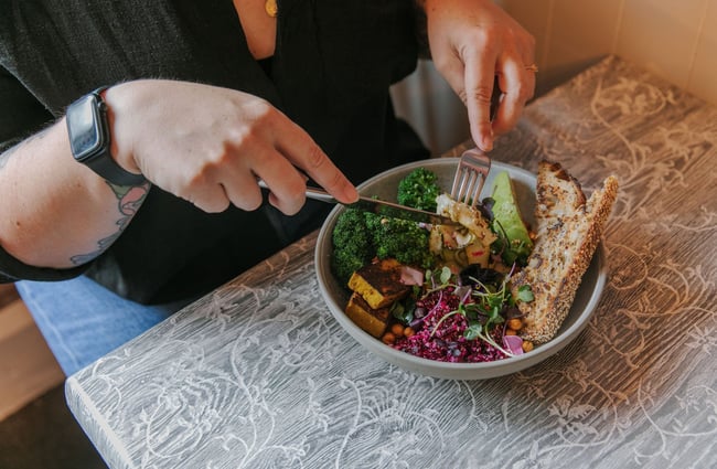 Close up of a person holding cutlery and cutting into a savoury Goodness Bowl with pickles, broccoli, avocado and multigrain bread.