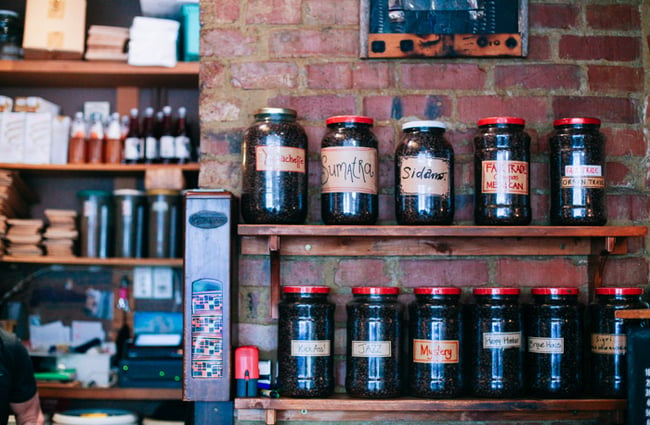 Jars of coffee beans on a wooden shelf against a brick wall.