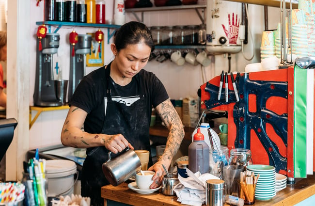 Barista with tattoos pouring milk into coffee behind cafe counter