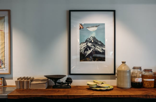 Framed artwork on the wall in the dining area above a wooden shelf with vintage scales, ceramic dishes and big jars of pickles sit in Muttonbird, Wānaka.