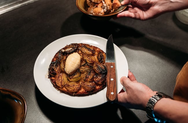 A staff member holding a plate of Muttonbird's onion tarte tatin with a scoop of parmesan custard on top of it, and a big, wooden-handled knife for serving.