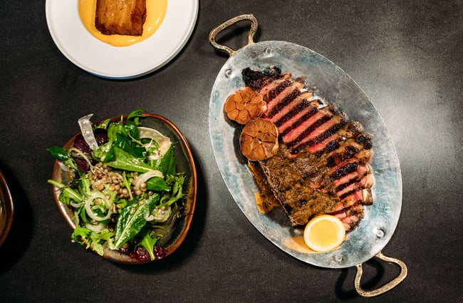 A silver dish of sliced red meat, garlic head halves and a lemon slice and a brown ceramic dish of salad greens sit on the pass at Muttonbird, Wānaka.