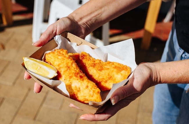 Outstretched hands holding a cardboard tray with two pieces of battered fish and a lemon slice.
