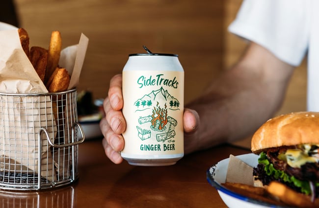 A hand holds a can of alcoholic ginger beer between a basket of chips and burger on a table at Red Star, Wānaka.