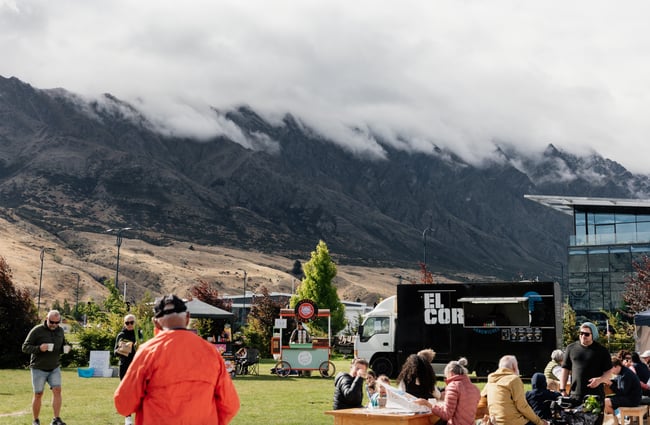 People sitting and standing around outside with mountains in the background.