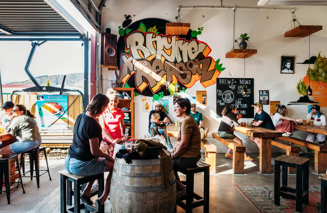 Groups of people sitting inside drinking beers in the sunshine in the high-ceilinged Rhyme and Reason taproom in Wānaka.
