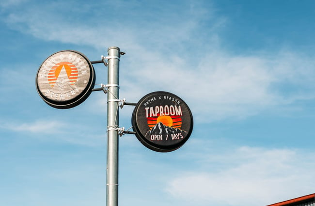 Two circular signs for the Brewery and Taproom mounted at the top of a tall pole outside Rhyme and Reason, Wānaka.
