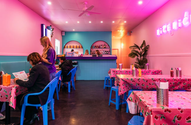 Staff serving customers at tables in the bright pink lit interior of Rice Rice Baby restaurant in Hamilton.