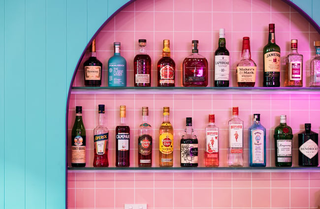 Liquor lined up on shelves in front of a pink tiled wall.