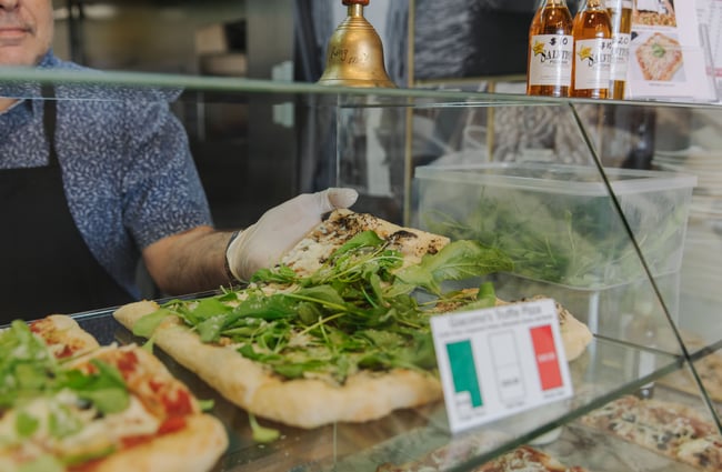 A worker wearing gloves grabbing a slice of pizza out of the glass display cabinet.