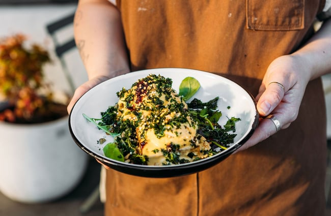 Staff member holding an enamel bowl of Scroggin's eggs Benedict with crispy kale on top.