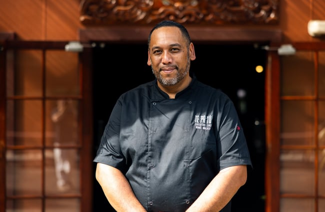 A man wearing a black chef's outfit smiling to camera.