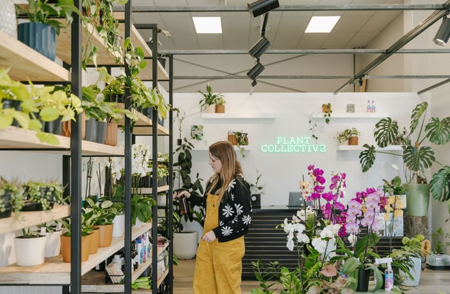 A women in bright yellow overalls looking at a wall of pot plants.