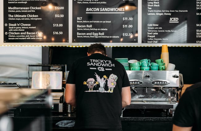 Close up of worker making coffee with Tricky's Sandwich Co logo on the back of his t-shirt