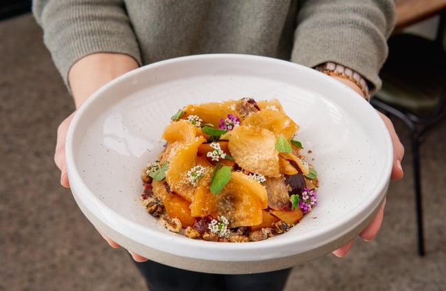 Close up of a staff member holding a white ceramic bowl of macerated fruit, mint leaves, hazelnuts and foraged flowers.