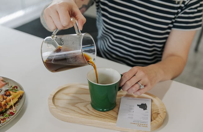 Someone pouring a filter coffee into a dark green cup on a wooden tray.