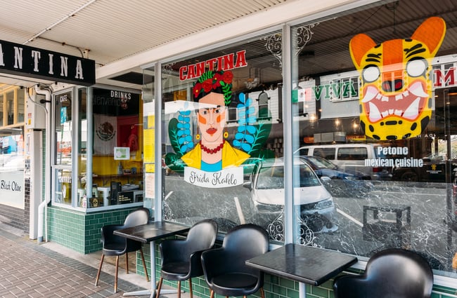 Exterior of Mexican restaurant with black dining chairs out front and illustrated window decals of Frida Kahlo and a tiger