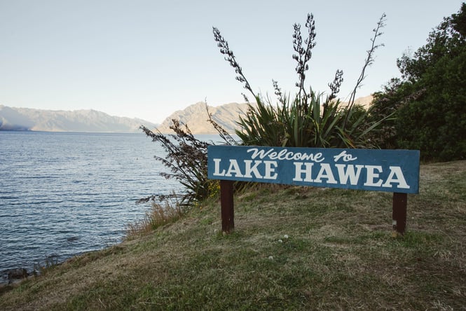 A sign saying 'Welcome to Lake Hawea' next to a lake.
