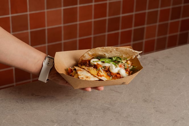 A hand holding a small cardboard box of tacos.