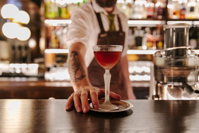 Bartender with tattooed arm holding out deep red cocktail in a frosted glass