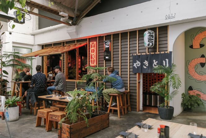 Diners sitting in outside area of Bar Yoku in The Welder space, surrounded by green plants, Japanese-inspired artwork and a red neon YOKU sign