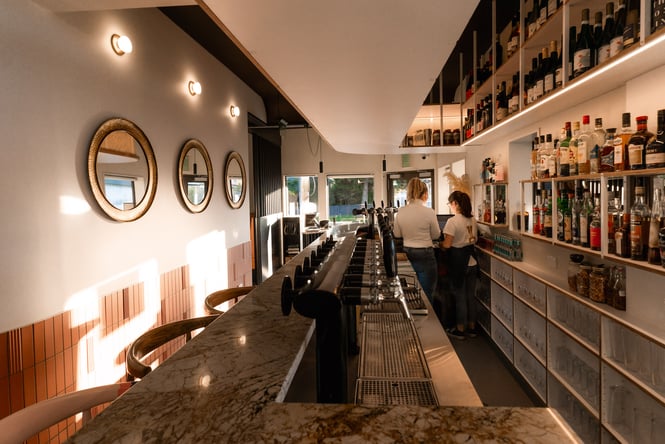 Overview of bar at the Birdwood with two women standing behind marbled counter and shelves of liquor and spirits behind them