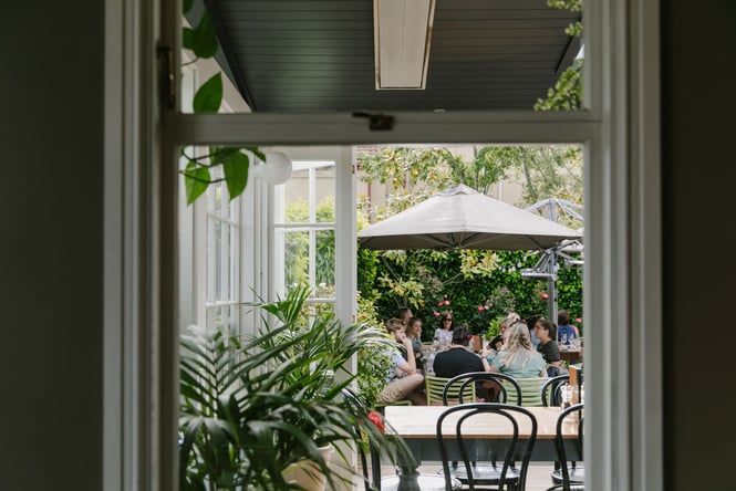 Looking through window out into the green courtyard at Dux Dine with diners eating and drinking underneath an umbrella
