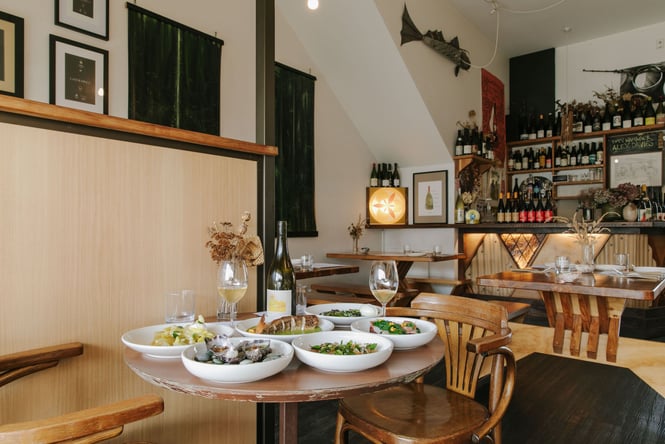 Round table filled with plates of seafood and vegetarian dishes with shelves of natural wine in the background behind the bar