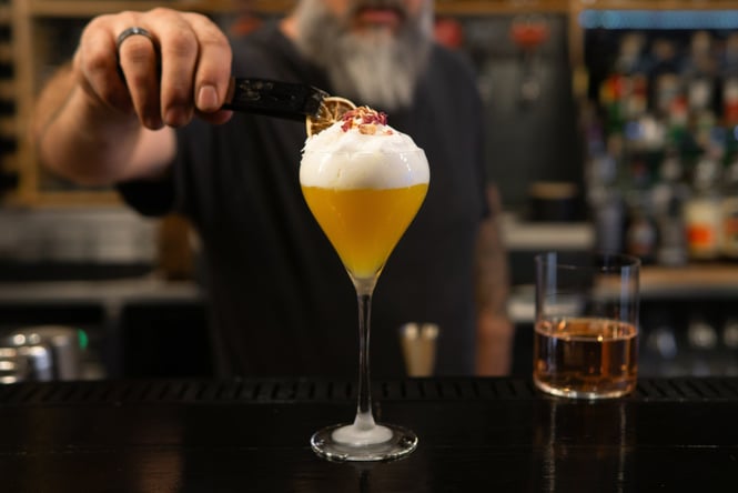 Bartender using tweezers to add dehydrated citrus garnish to foamy pink cocktail
