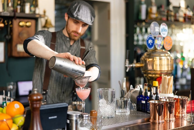 Bartender in old-fashioned newsboy cap pouring pink cocktail into glass at OGB bar