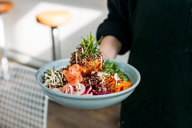 Close up of a waiter holding a shallow bowl of salad with sliced radish, grated carrot, pickled ginger and more colourful ingredients topped with battered sticky tofu and coriander.