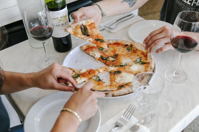 People enjoying red wine and picking up a slice of Margherita pizza at a restaurant