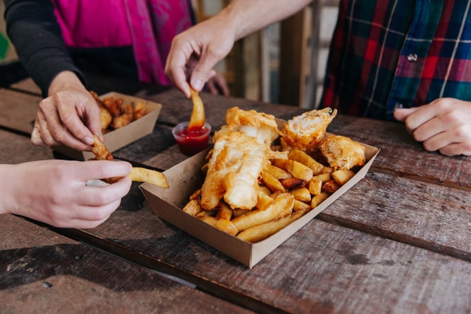 Three people sitting around a wooden table holding hot chips next to a box of deep fried fish.