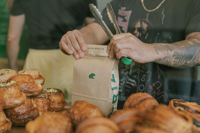 Staff member holding tongs while filling Grizzly brown paper bag next to morning buns and croissants