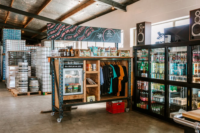 Corner of Urbanaut Brewery with fridges full fo beer and DIY deli platters, packets of chips on the shelf and branded merch on racks