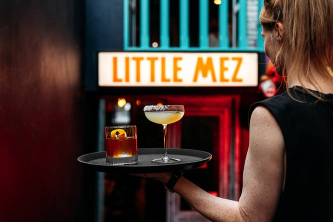 Bartender holding black tray with two cocktails under illuminated Little Mez sign