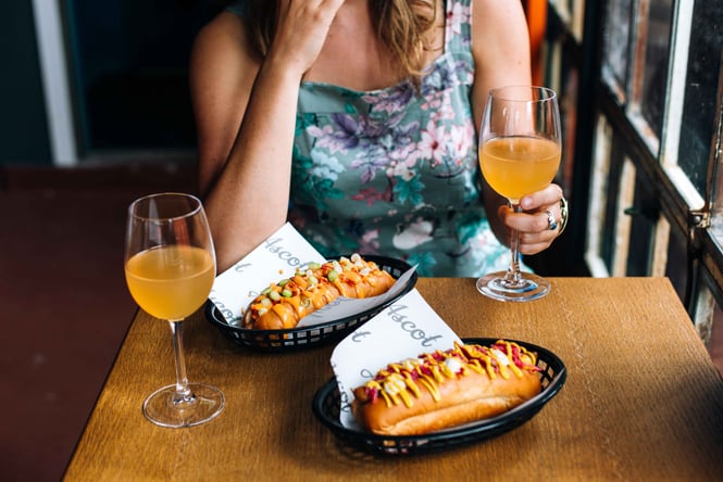 A woman sitting next to two glasses of wine and two hot dogs.