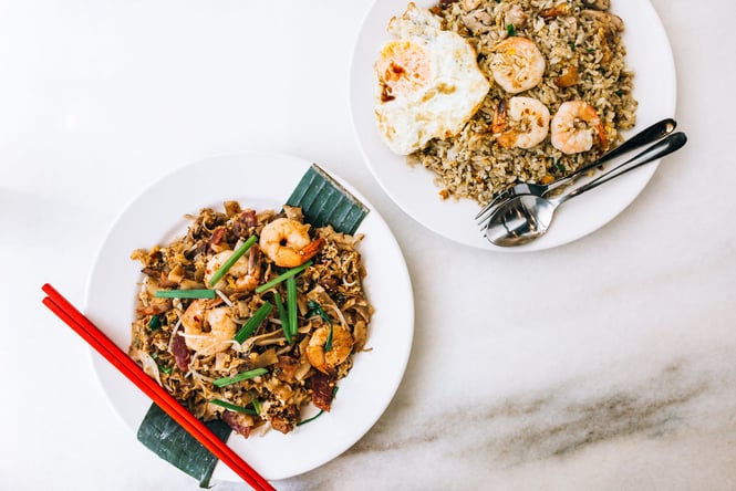 A flatlay of a plate of noodles and a plate of rice on a white table.