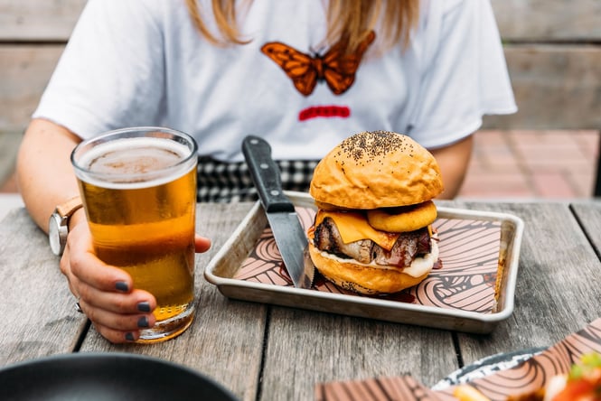 A person holds a full pint of beer on a table beside a burger stacked with bacon and onion rings on a tray with a big knife, at Salt and Wood.