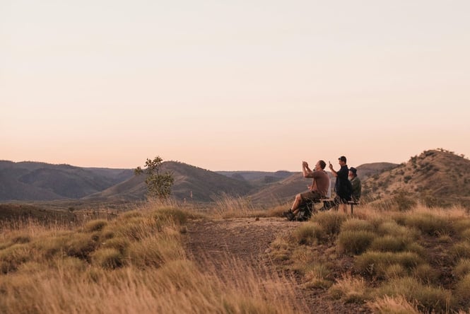 Hikers sit on a bench on a tussocky hill, taking photos of the golden light.
