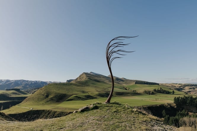 A metal sculpture of a tree blowing in the wind on a hilltop in Canterbury, overlooking other grassy hills.